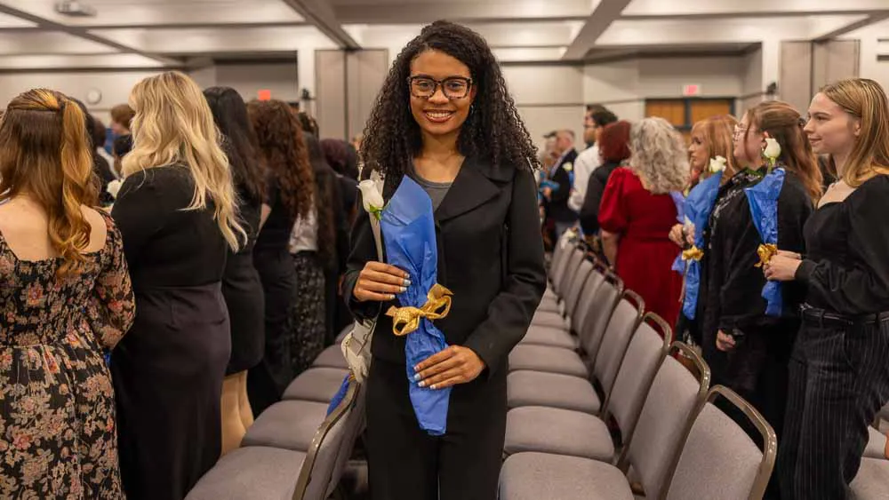 A smiling Wake Tech student holds a white rose wrapped in blue paper during the Phi Theta Kappa induction ceremony.